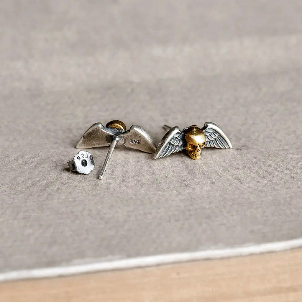 Ear studs-örhängen av äkta silver föreställande en guldfärgad dödskalle med vingar