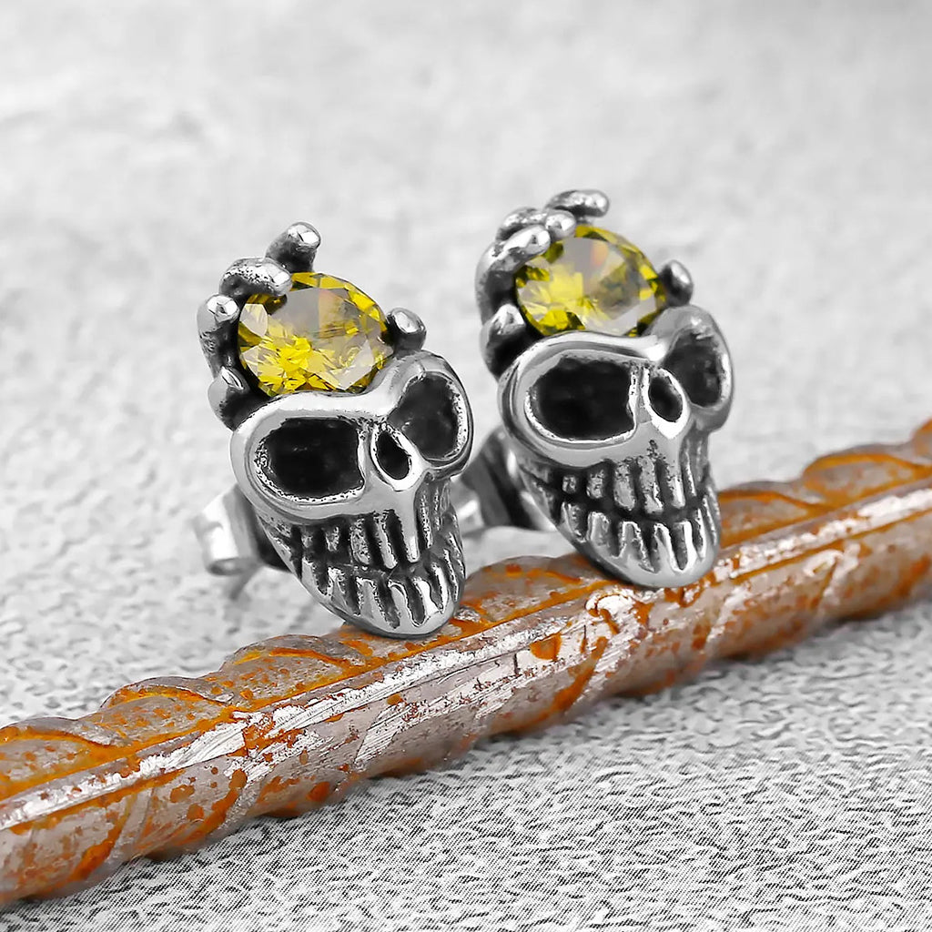 Silverfärgat stiftörhänge som visar en dödskalle med en skeletthand hållande en gul zirkon